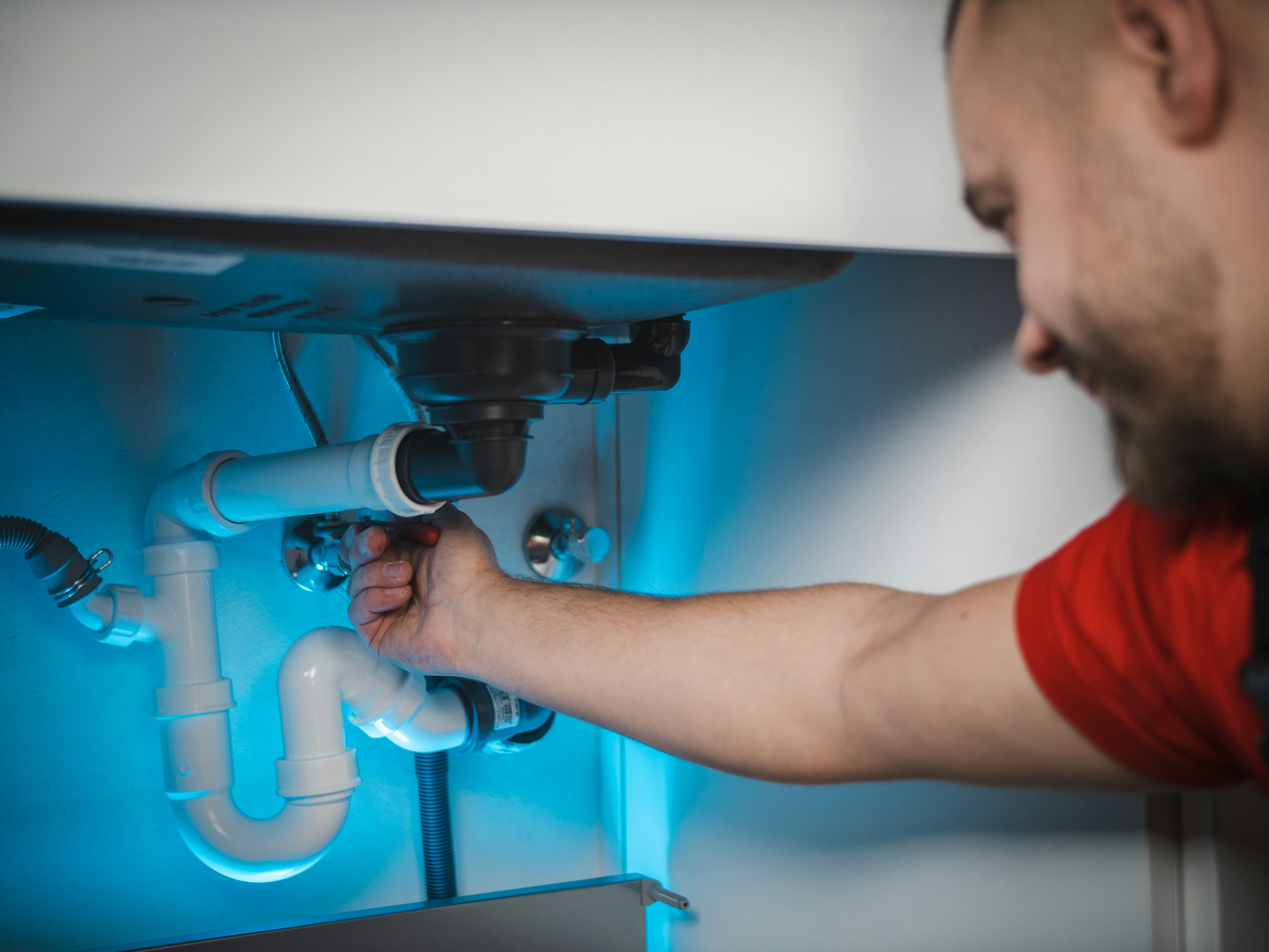 Plumber in a red T-shirt reaching under a kitchen sink to tighten a drain pipe