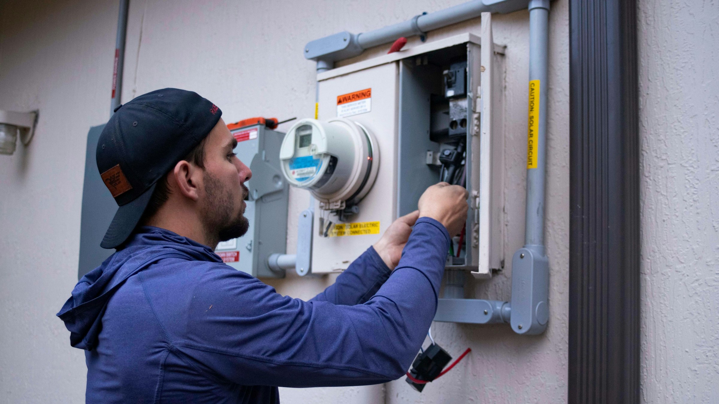 Electrician in a blue hoodie wiring a meter panel on an exterior wall
